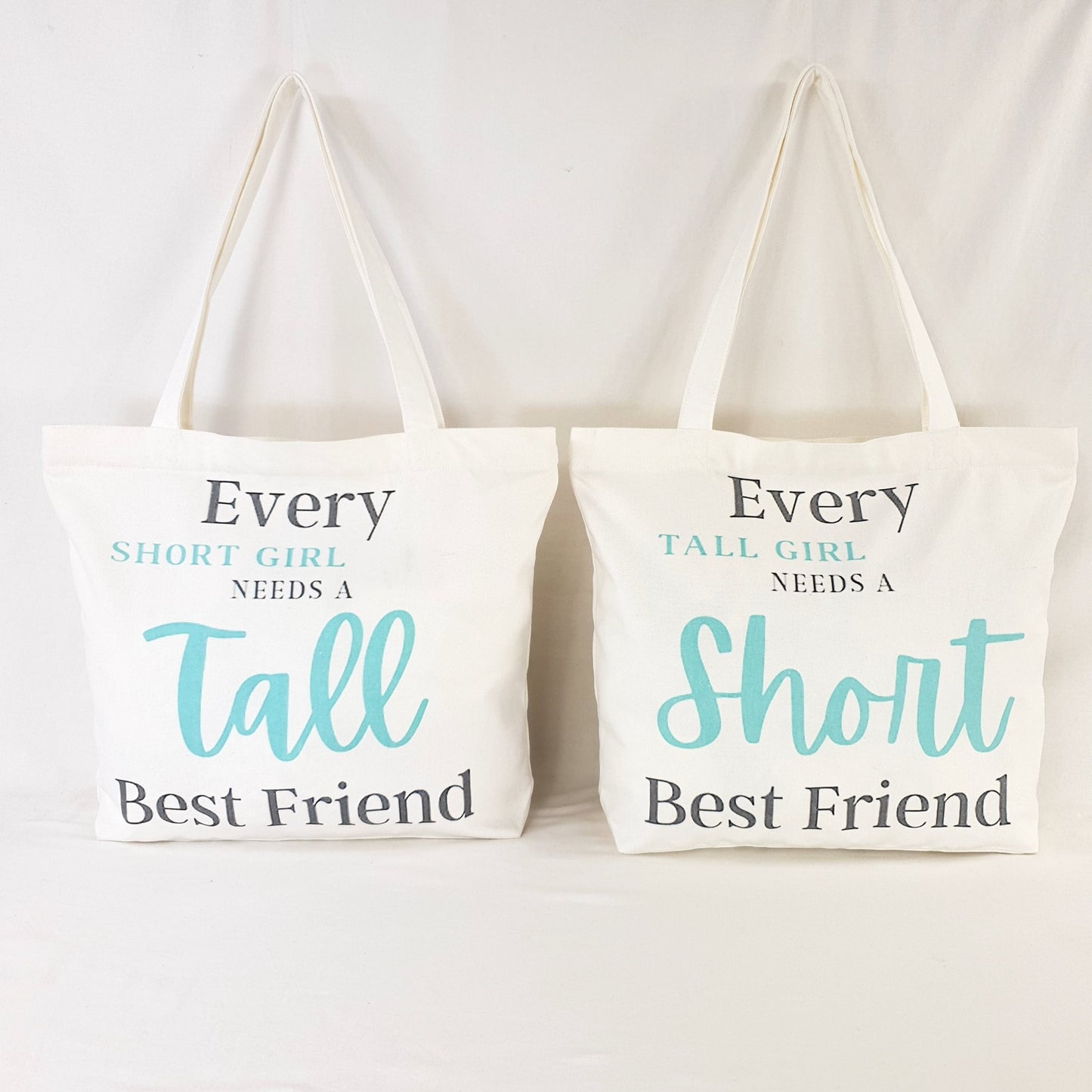 Tall Friend and Short Friend bags, one bag print text reads : "Every Short girl needs a tall best friend" and the words "short girl" and "Tall" is in Teal color and rest of writing is in dark grey. the other bag print reads: "Every tall girl needs a short best friend" colouring of the print text is the same as the other bag.  Bag itself is white with shoulder straps, zipper on the top