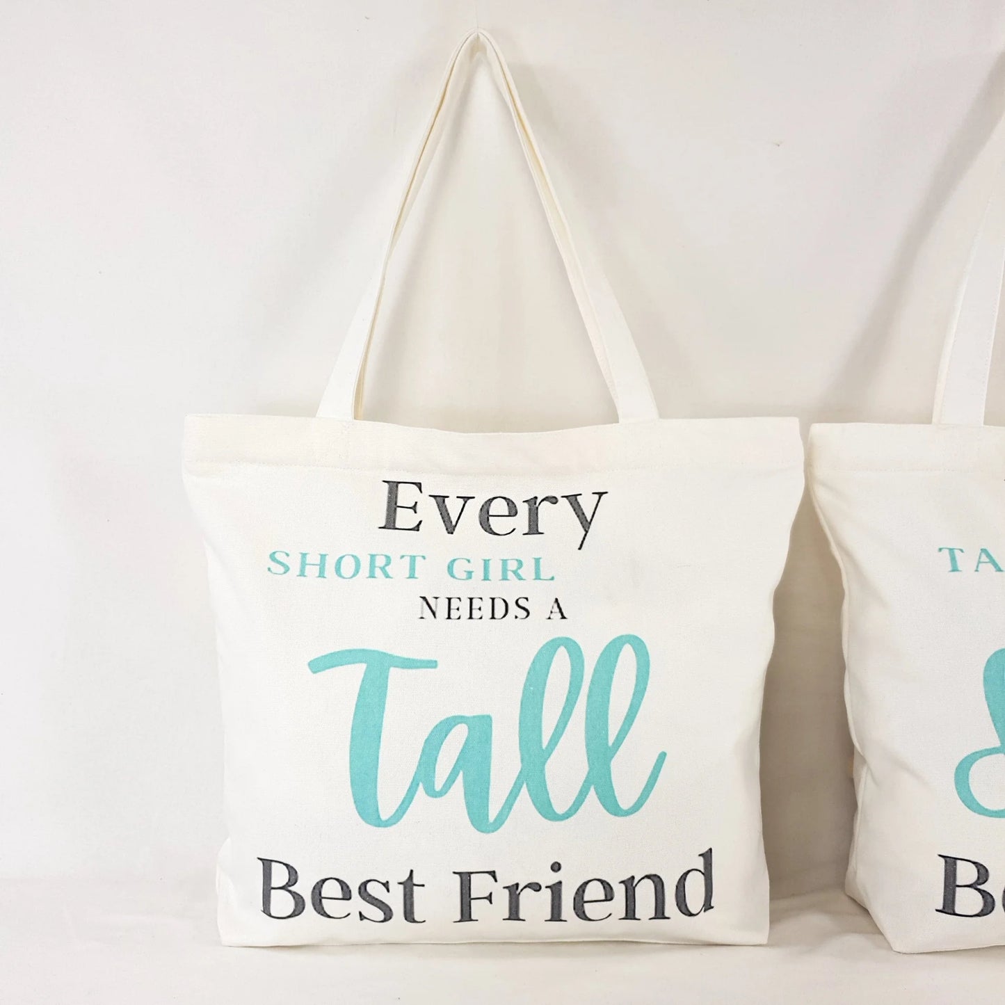 printed text reads : "Every Short girl needs a tall best friend" and the words "short girl" and "Tall" is in Teal color and rest of writing is in dark grey.  Bag itself is white with shoulder straps, zipper on the top